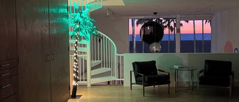 A spacious room with a staircase, a view of the sunset through a window, and a seating area with chairs and a table.