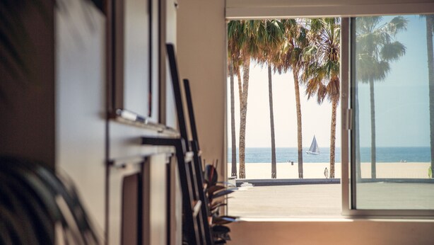 A room with a large window overlooking a beach with palm trees and a sailboat.