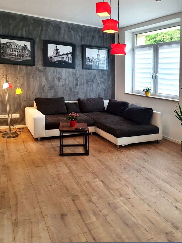 A modern living room with a sectional sofa, a coffee table, and a floor lamp. There are framed pictures on the wall and a window with blinds. - Modern and spacious apartment, with separate access (Dohna)