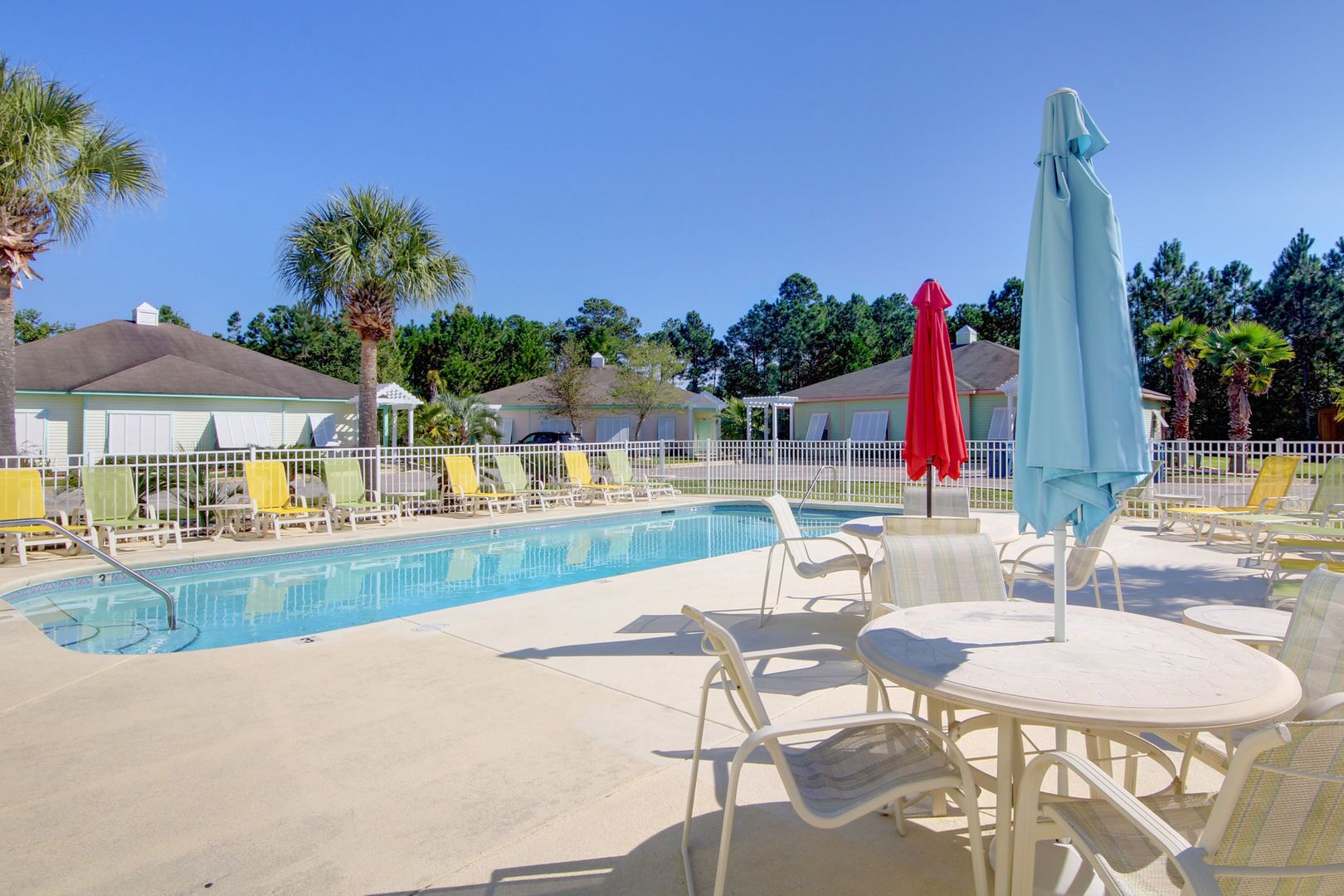 A pool area with lounge chairs and umbrellas, surrounded by a white fence and palm trees.