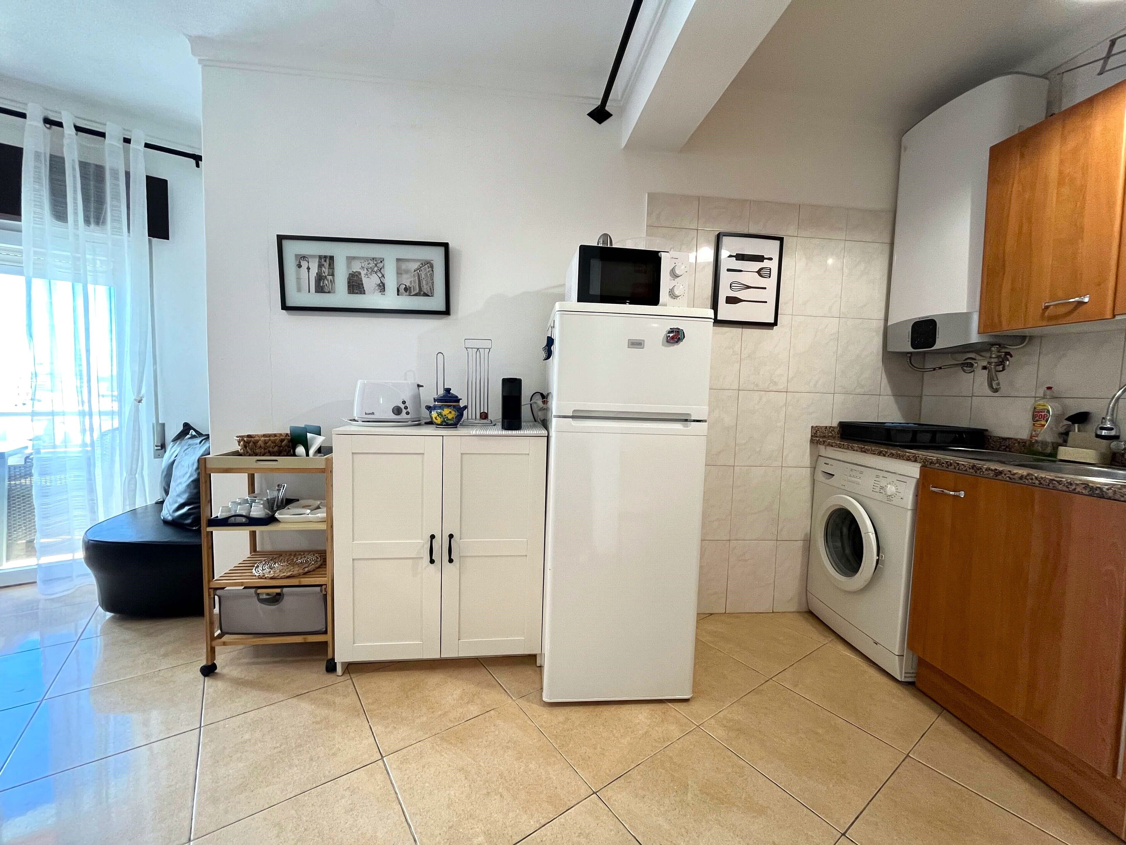 A compact kitchen with a refrigerator, microwave, and washing machine. There is a small dining area with a table and chairs.