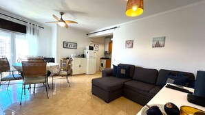Un soggiorno moderno con un divano componibile, un'area pranzo con sedie in vimini, una cucina con elettrodomestici bianchi e una ventola a soffitto.