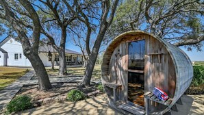 Una estructura rústica con forma de barril de madera, con un banco en su interior, rodeada de árboles y una casa al fondo.