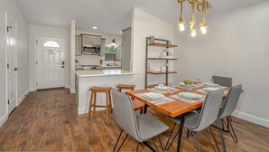 Un coin repas avec une table en bois dressée pour quatre personnes, entourée de chaises à revêtement gris. On aperçoit, au fond, une cuisine équipée d’appareils électroménagers modernes.