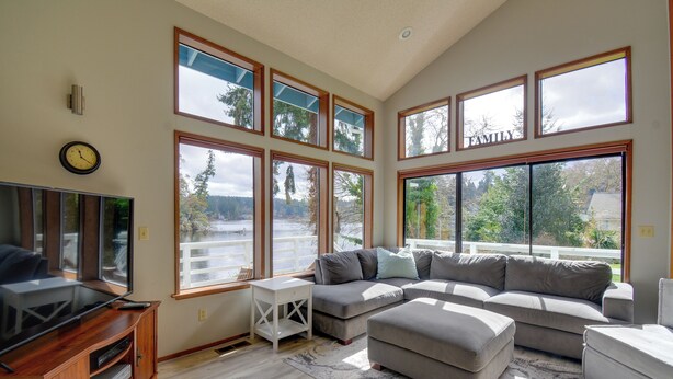 A spacious living room with a large sectional sofa, a flat-screen TV on a wooden stand, and multiple large windows offering views of a lake and trees. - The Swedish Fish: Getaway on Lake Saint Clair! (Olympia)