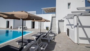 Un área junto a la piscina con sillas de descanso y sombrillas, una piscina azul cristalina y un edificio blanco con una escalera.