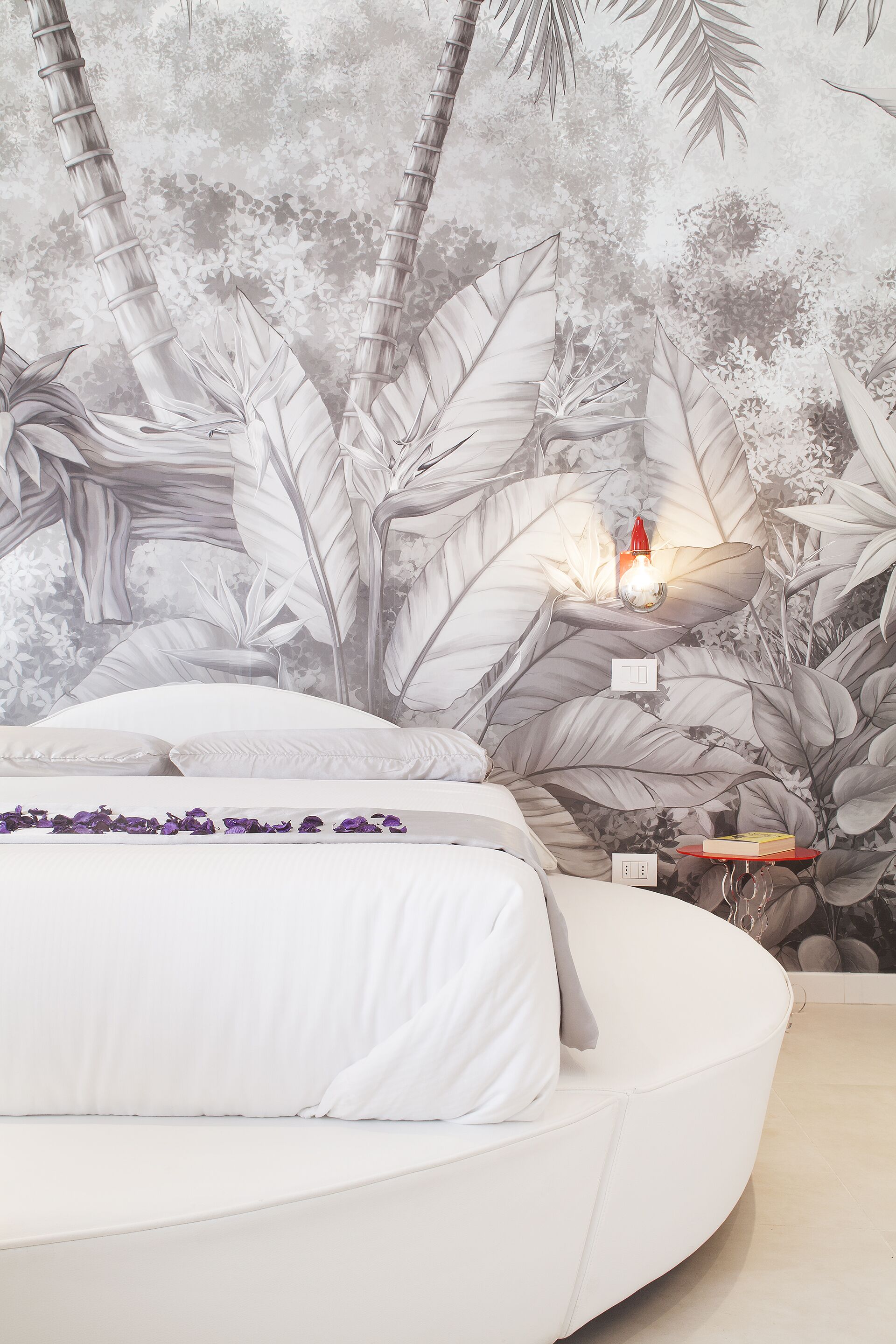 A bedroom with a white bed, a nightstand with a red lamp, and a wall mural of tropical leaves.