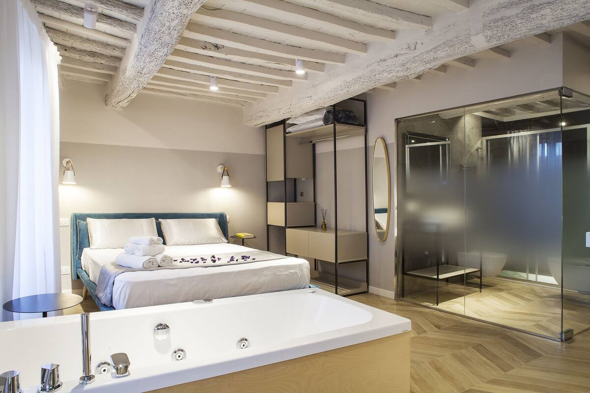 A modern hotel room with a large bed, a separate bathroom with a glass shower, and a wooden floor.