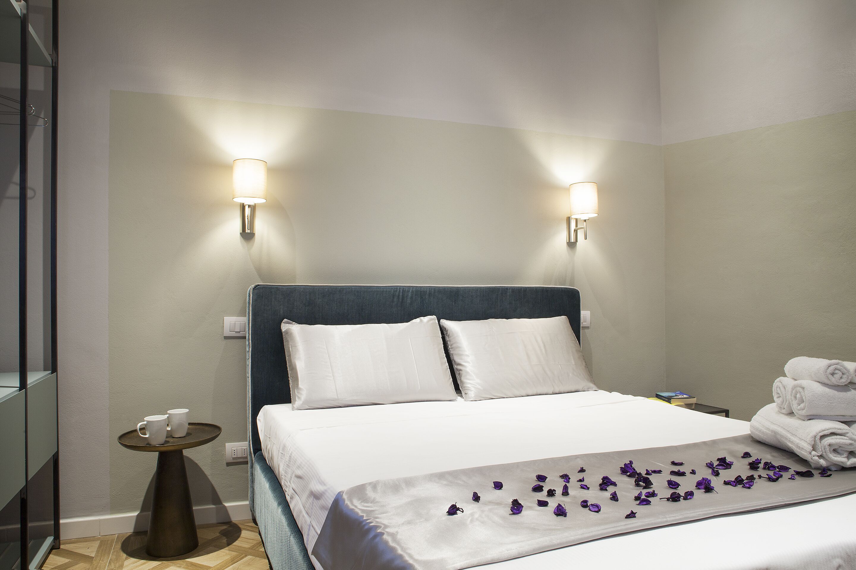 A hotel room with a bed, two wall-mounted lights, a small side table, and a neatly made bed with towels.