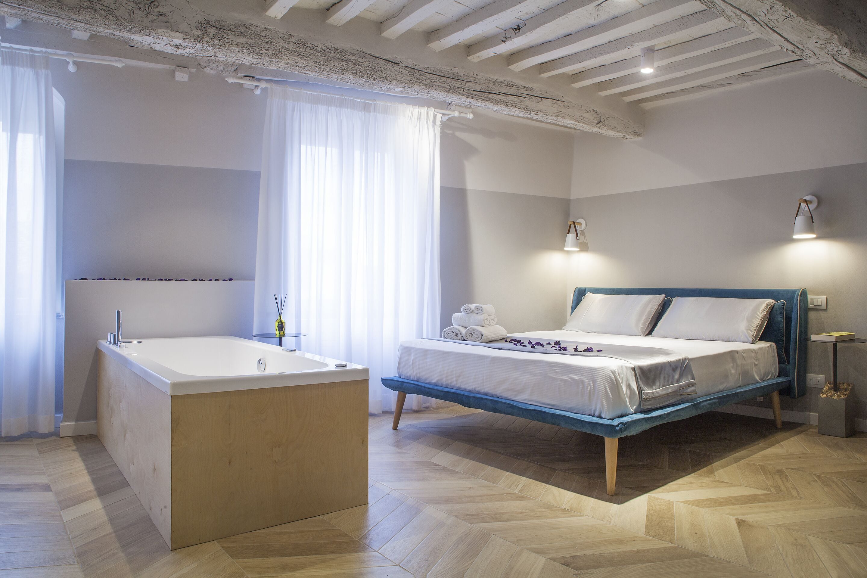 A modern bedroom with a wooden floor, a large bathtub, a bed with blue headboard, and two bedside lamps.