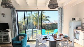 Un salon moderne doté d’une porte-fenêtre coulissante donnant sur une terrasse, d’un coin repas avec des chaises blanches, d’une cuisine avec des appareils électroménagers au style rétro et d’un téléviseur posé sur un meuble.