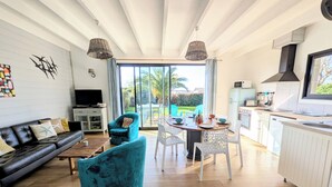 Un salon moderne avec un canapé en cuir noir, un fauteuil turquoise et une table basse en bois. On y trouve également un coin repas avec une table et des chaises blanches, ainsi qu’une cuisine avec des meubles blancs et une hotte noire.