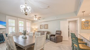 Un vaste espace de vie comprenant une table à manger, des chaises, un canapé et un fauteuil inclinable. On y trouve également un ventilateur de plafond, un lustre et un téléviseur fixé au mur.