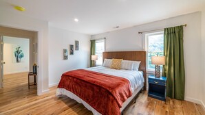 A bedroom with a wooden floor, a bed with a red blanket, a nightstand with a lamp, and a window with green curtains. - East Virginia Blues Condo, Walk to Beach & Shops (Cape Charles)