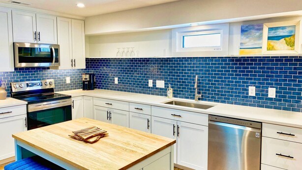 A modern kitchen with white cabinets, a blue-tiled backsplash, stainless steel appliances, and a wooden island with stools.