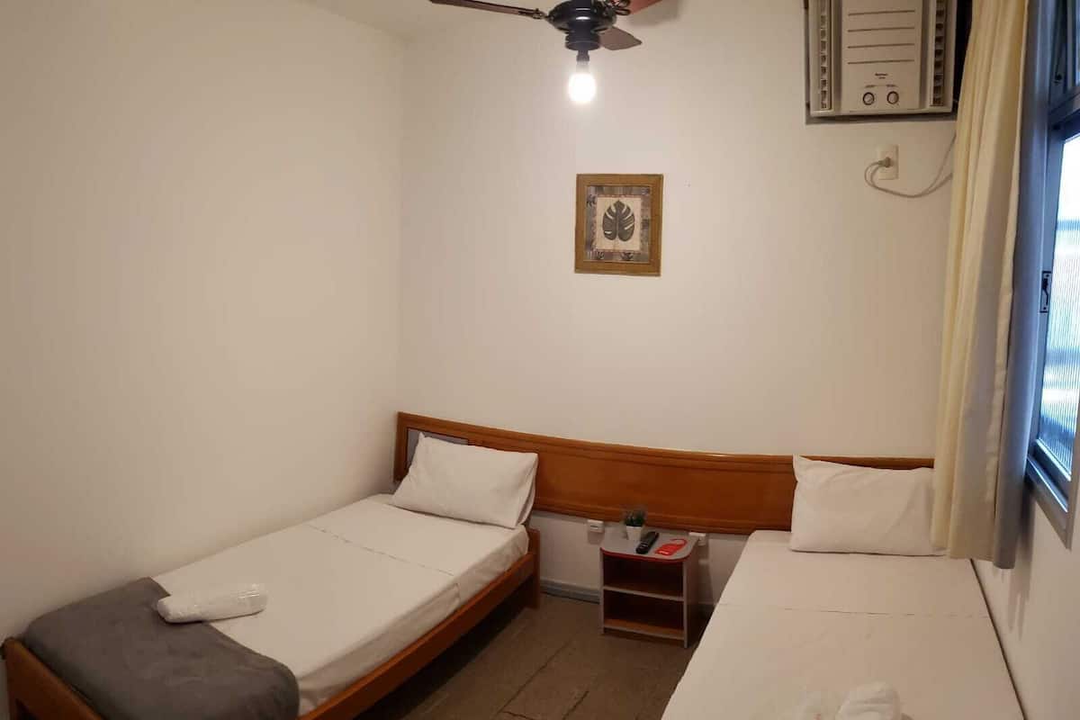 Una habitación pequeña y limpia con dos camas individuales, un ventilador de techo, una mesita de noche con control remoto y una ventana con cortinas.