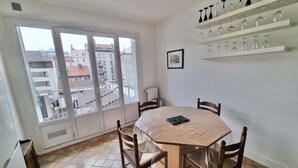 A dining area with a round table, chairs, a window with a view of buildings, and a shelf with glassware. - #CX |Chez Faffa 𓅂| Spacious T5 | Train station | (Grenoble)