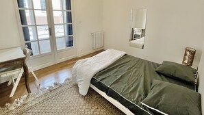 A bedroom with a bed, a desk, a chair, a radiator, and a window with a view of buildings outside. - #CX |Chez Faffa 𓅂| Spacious T5 | Train station | (Grenoble)