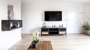 Una sala moderna con una televisión de pantalla plana montada en la pared, una mesa de centro de madera con una orquídea en maceta, un mueble multimedia negro con objetos decorativos y una puerta blanca.