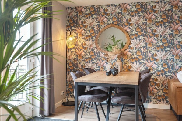A dining area with a wooden table, dark chairs, a floral wallpaper, a mirror, and a plant.