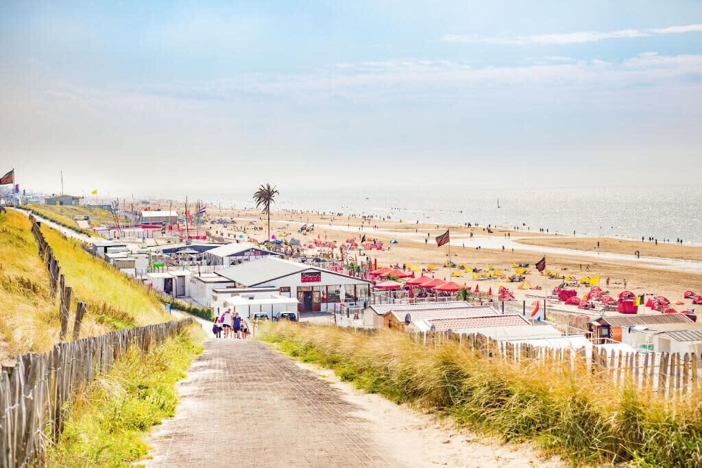 Eine Küstenlandschaft mit sandigem Strand, einem Holenzaun und einem gepflasterten Weg, der zu einem Gebäude mit rotem Dach führt.