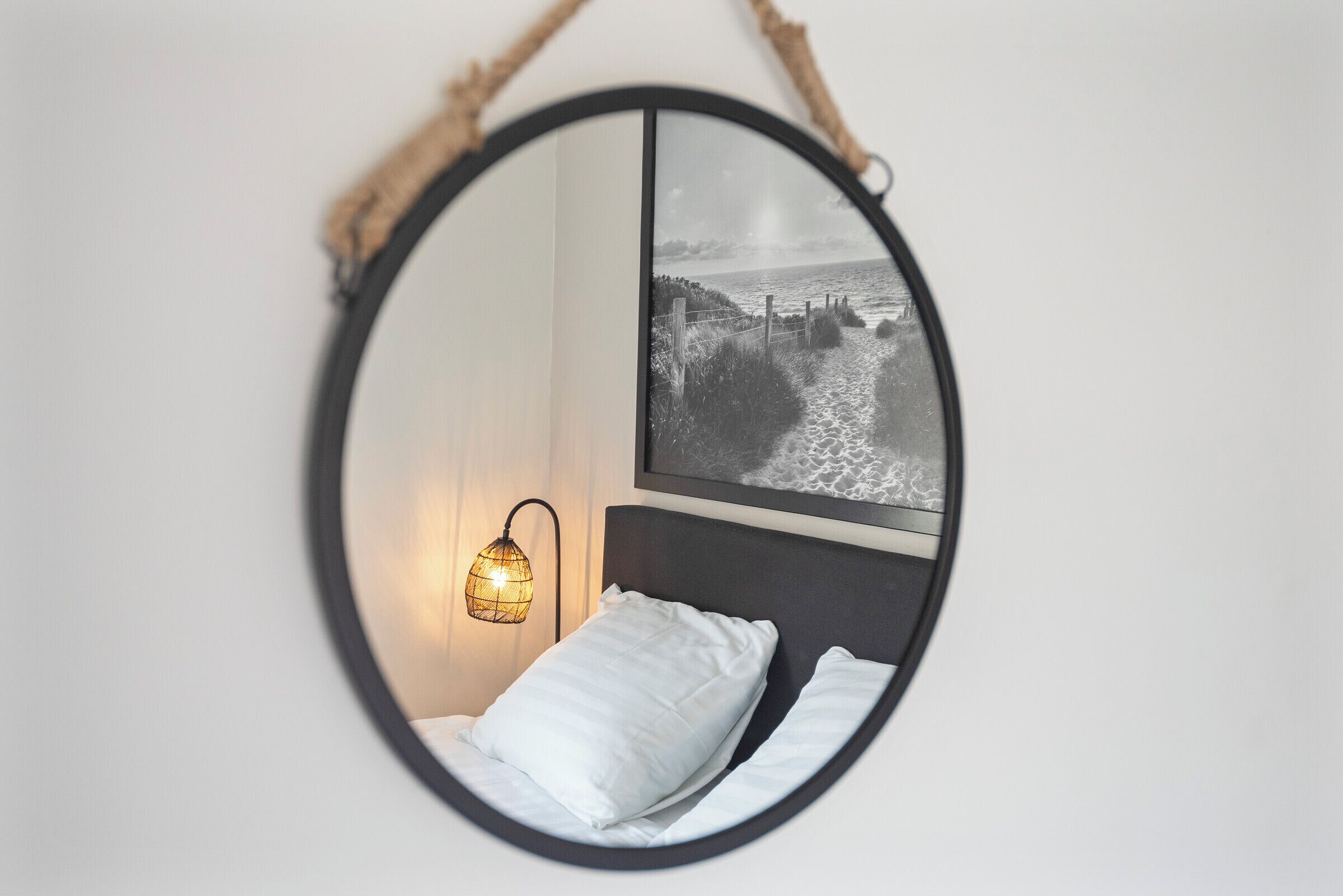 Un miroir rond avec un cadre noir, qui reflète un lit avec une literie blanche et une lampe fixée au mur.