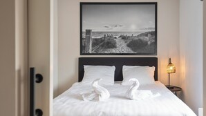 Un lit bien fait, avec du linge de lit blanc et deux oreillers en forme de cygne, une photographie en noir et blanc au-dessus du lit et une lampe de chevet sur le côté droit.