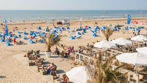 Une plage parsemée de nombreuses tentes bleues et de parasols blancs, avec une tour de surveillance et une zone de plage sablonneuse équipée de chaises longues et de tables.