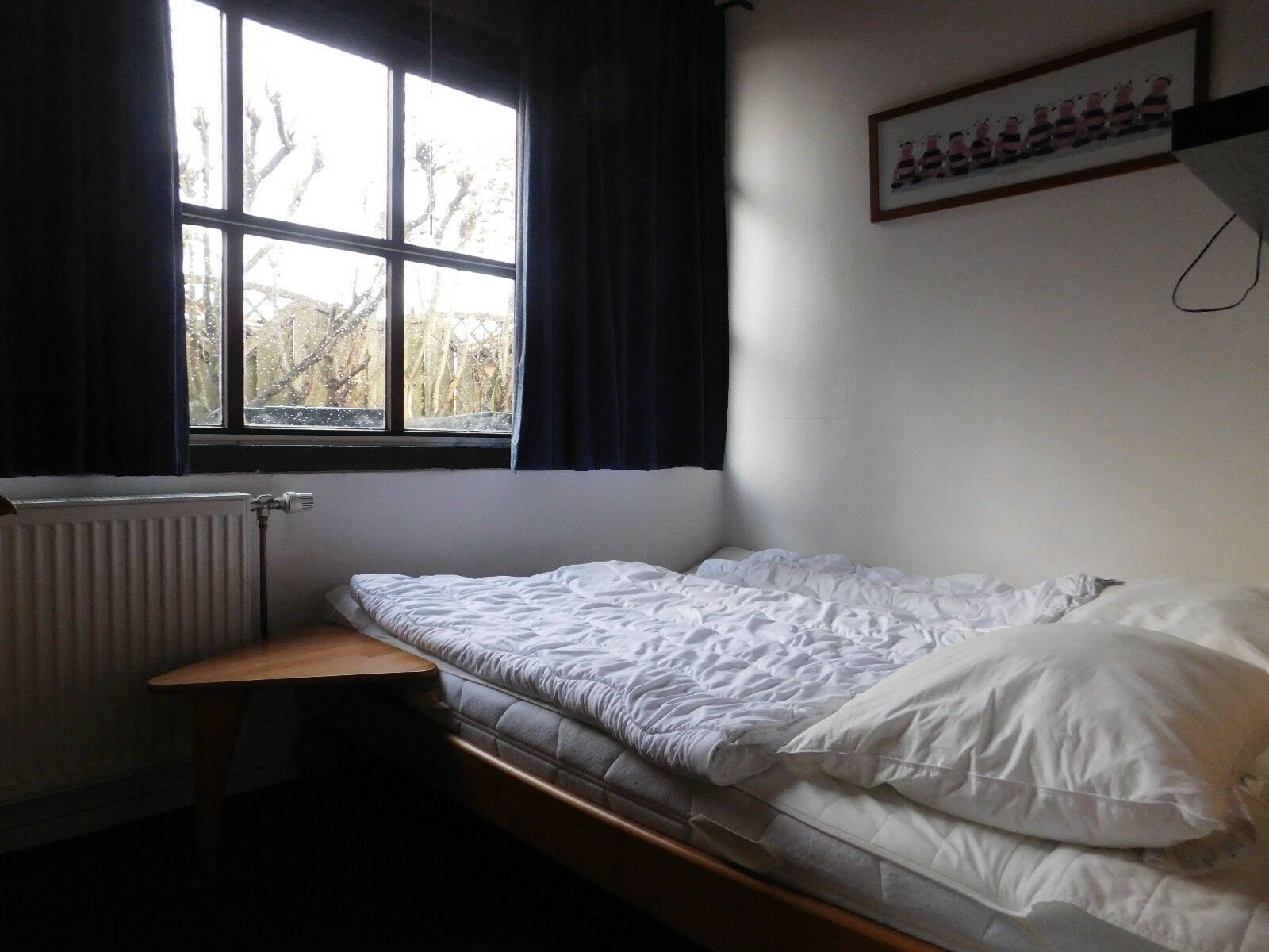 A small, single-bed room with a window, a radiator, and a framed picture on the wall.
