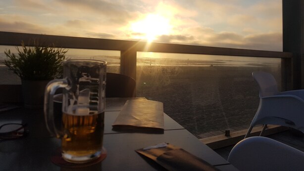 Un tavolo con un bicchiere di birra, un libro e una sedia, con vista su un tramonto sull'acqua.