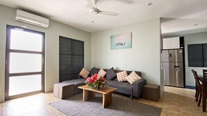 Ett modernt vardagsrum med en grå soffa, ett soffbord i trä och en grå matta. Här finns också en köksdel med ett kylskåp i rostfritt stål och ett matbord med stolar.
