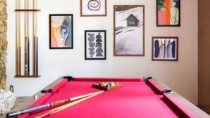 A pool table with a red felt surface, surrounded by framed artworks on the wall, and a set of pool cues to the left. - Lakeshore Villa (Incline Village)