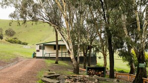 A rustic cabin with a dirt path leading to it, surrounded by trees and a picnic area with benches and a firewood stack. - Strath Creek Lodge at The Village Green (Strath Creek)