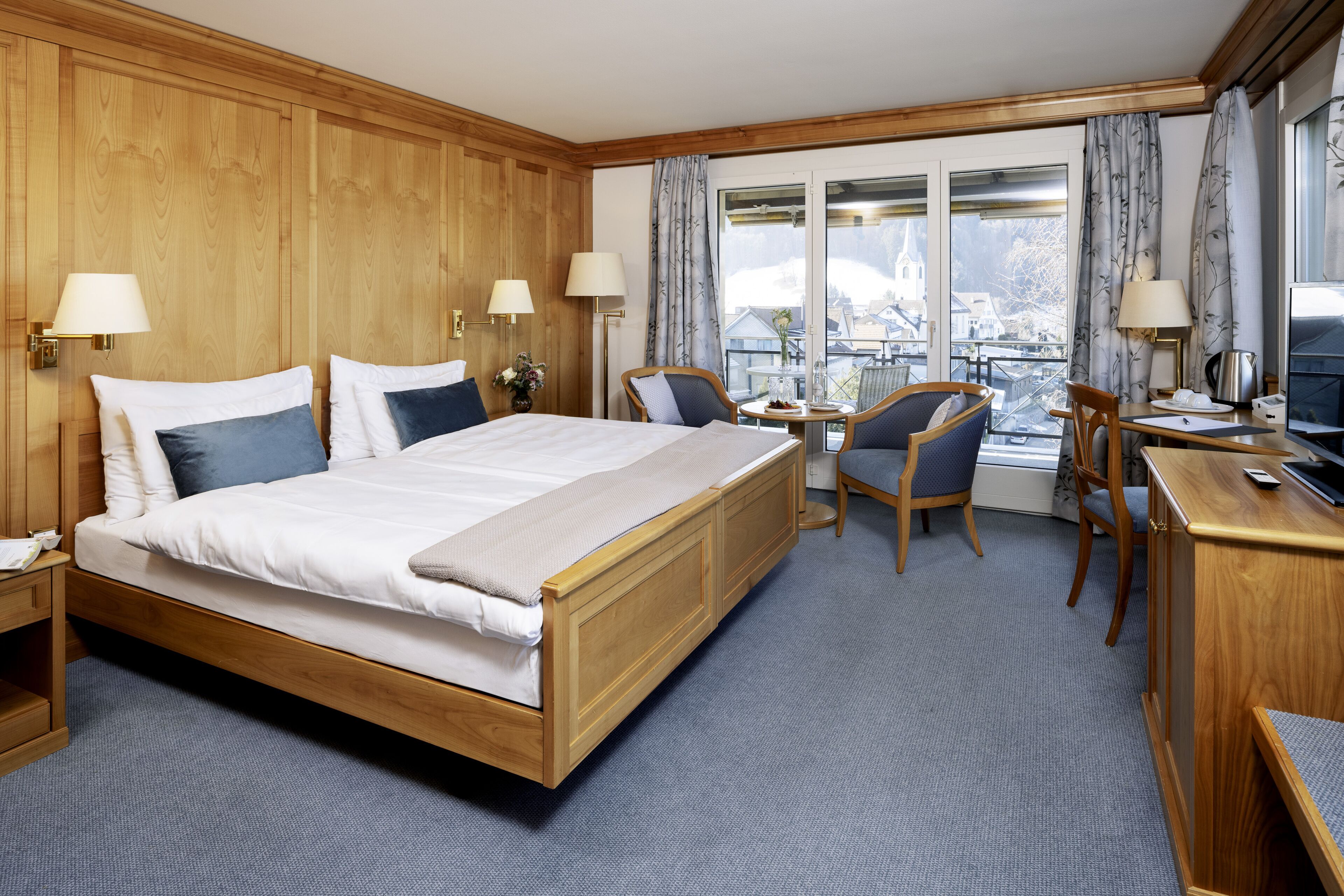 Une chambre d’hôtel avec un lit en bois, deux chaises, un bureau et une vue sur un paysage enneigé, visible à travers de grandes fenêtres.