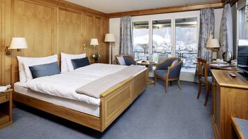 Une chambre d’hôtel avec un lit en bois, deux chaises, un bureau et une vue sur un paysage enneigé, visible à travers de grandes fenêtres.