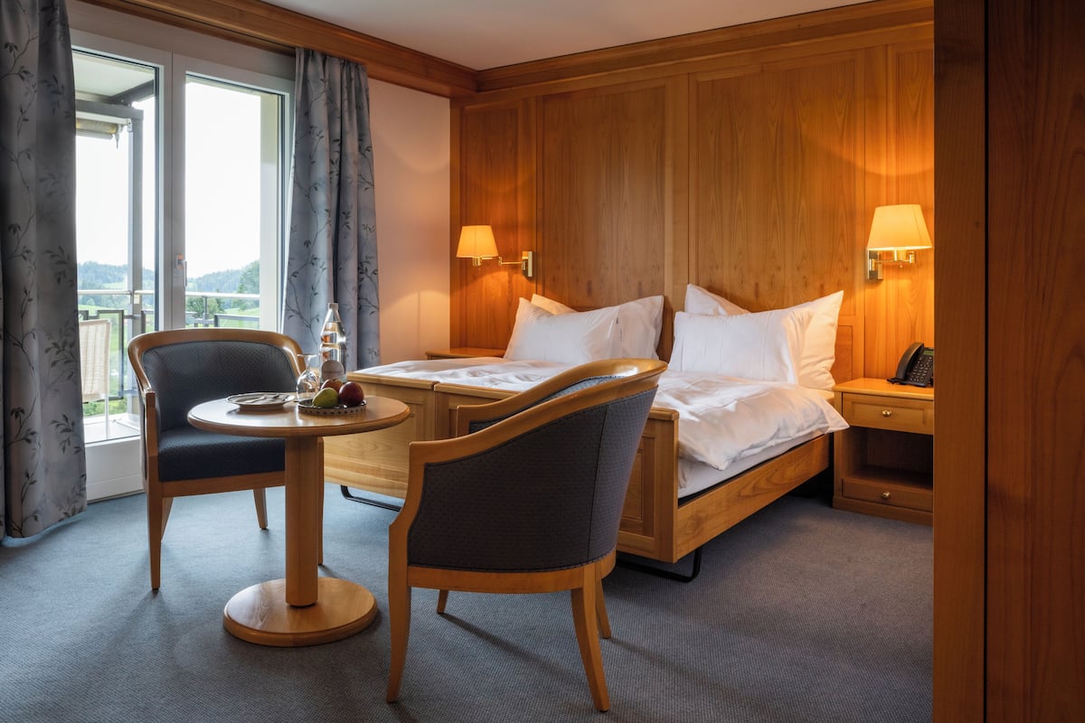Ein Hotelzimmer mit einem Bett, zwei Stühlen, einem kleinen Tisch mit Obst, einer Lampe und Blick ins Freie.