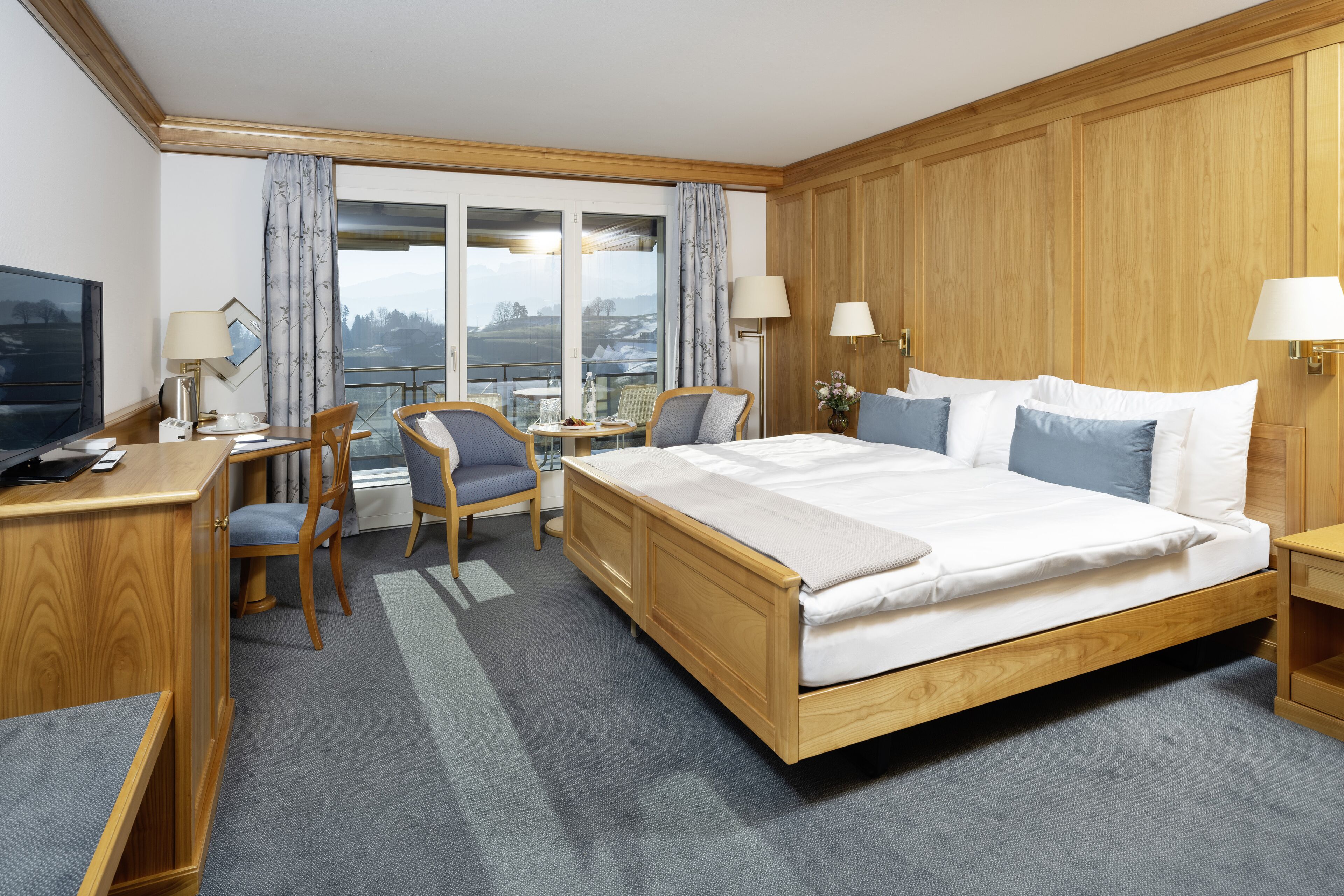 Ein Hotelzimmer mit einem großen Bett, einem Holzkopfende, einem Schreibtisch mit Stuhl, einem kleinen Tisch und einem Fernseher.