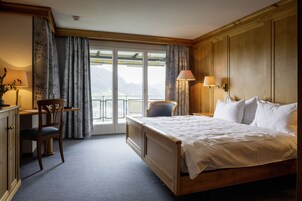 Habitación de hotel con cama de madera, un escritorio con silla, balcón con cortinas y vistas a las montañas.