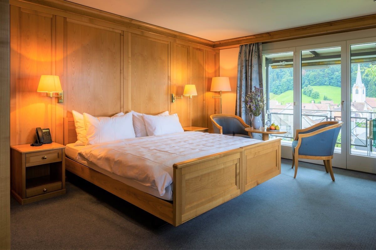 Ein Hotelzimmer mit einem großen Bett, einem Holzkopfstück, zwei Nachttischlampen, einem Sessel und Blick auf eine grüne Landschaft durch das Fenster.