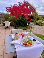 Mesa de comedor al aire libre con diversos platos y bebidas, rodeada de vegetación exuberante y un árbol de hojas rojas.