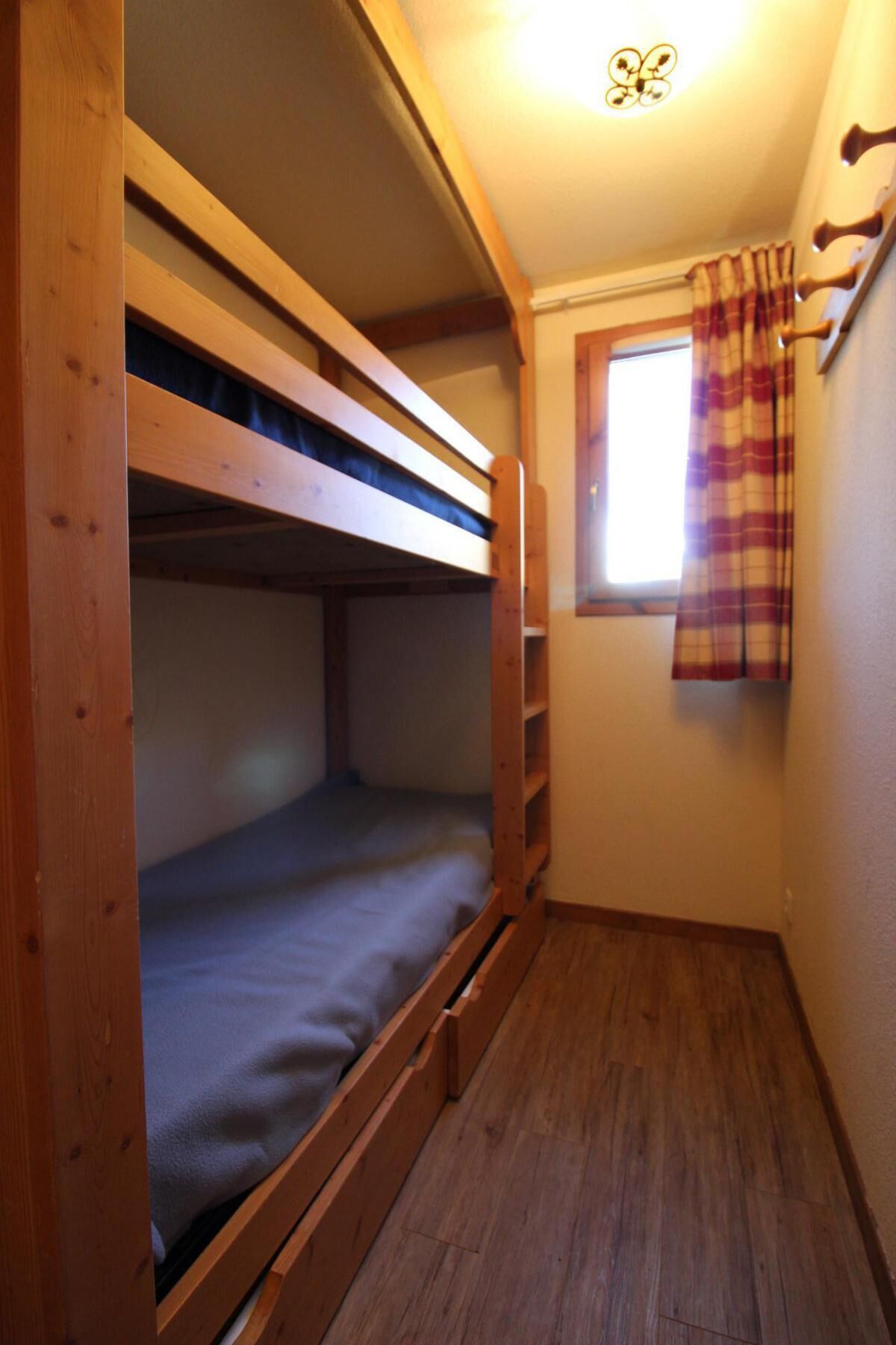 A small, narrow room with a bunk bed and a window with plaid curtains.
