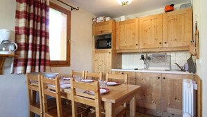 A wooden dining table with chairs, a kitchen with wooden cabinets, a microwave, and a window with curtains. - 150m from shops, Ground floor, view mountain, fitness, swimming pool, balcony, tv, ski locker, 38m² (Lanslebourg-Mont-Cenis)