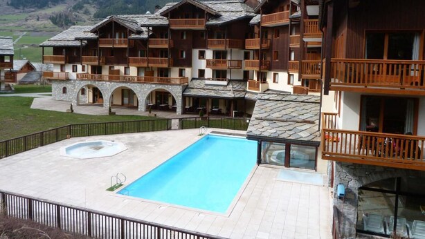 A hotel with multiple wooden balconies, a swimming pool, and a mountainous backdrop. - 150m from shops, Ground floor, view mountain, fitness, swimming pool, balcony, tv, ski locker, 38m² (Lanslebourg-Mont-Cenis)