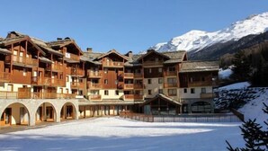 A multi-story wooden building with balconies, set against a snowy mountain backdrop. - 150m from shops, Ground floor, view mountain, fitness, swimming pool, balcony, tv, ski locker, 38m² (Lanslebourg-Mont-Cenis)