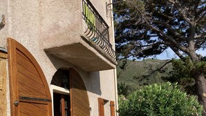 Ein rustikales Gebäude mit Natursteinfassade, Holztüren und einem Balkon mit schmiedeeisernem Geländer. Auf der Terrasse befinden sich Sitzgelegenheiten und Kübelpflanzen.