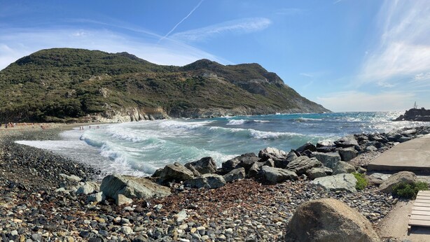 Ein felsiger Strand mit brechenden Wellen, ein hölzerner Steg und eine gebirgige Kulisse.