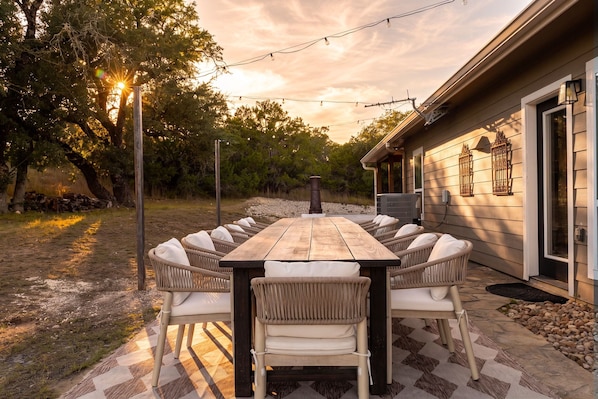 An outdoor dining area with a wooden table and wicker chairs, surrounded by a stone wall and trees, with a house in the background.