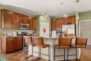 A kitchen with wooden cabinets, a stainless steel refrigerator, a microwave, and a bar area with leather chairs.