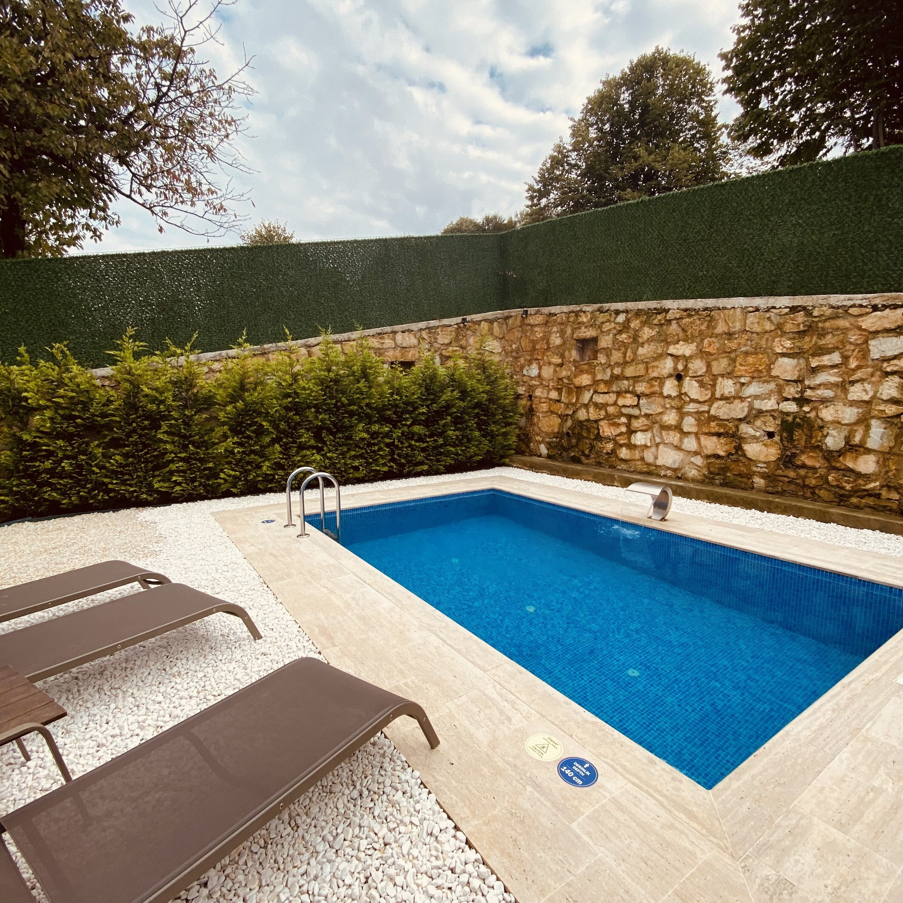 Una piscina rectangular con un muro de piedra y un seto verde al fondo.