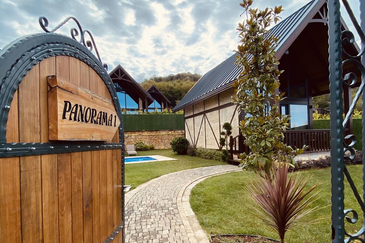 Une porte en bois portant l’inscription « PANORAMA 1 » mène à un bâtiment doté d’un toit en pente, avec une piscine en arrière-plan.